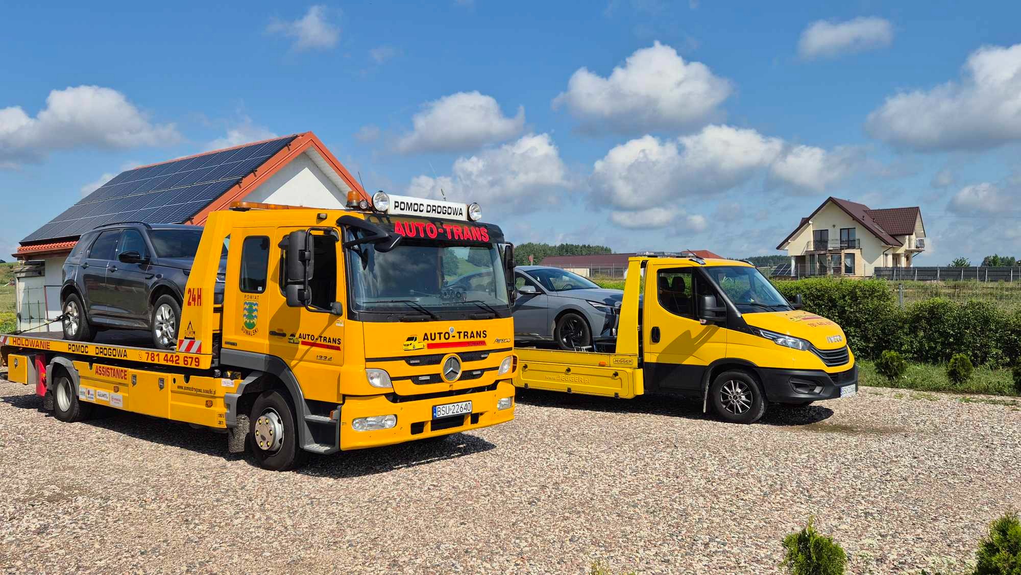 Dwa żółte pojazdy pomocy drogowej z załadowanymi samochodami osobowymi na żwirowym podjeździe przed domami z panelami słonecznymi na dachu, błękitne niebo z białymi chmurami w tle.