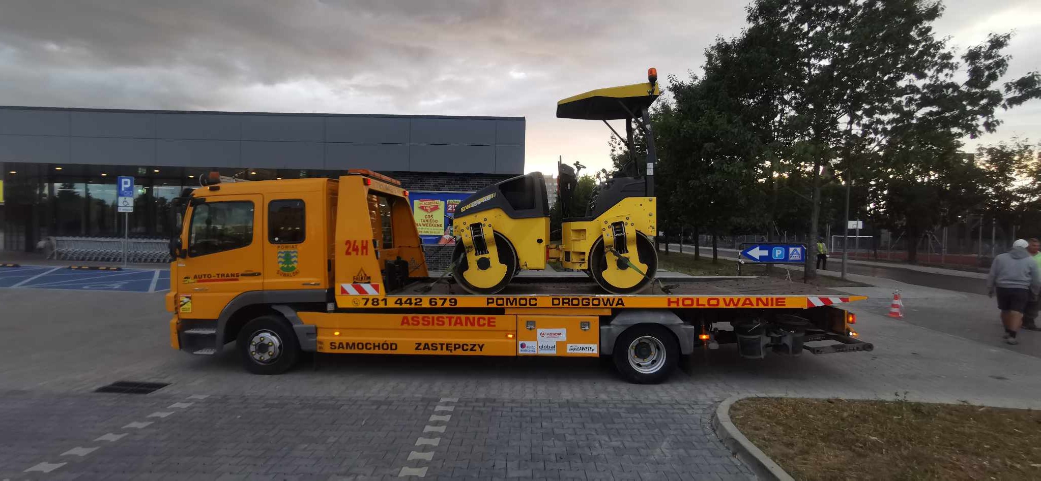 Żółta laweta z pomarańczowymi oznaczeniami i napisem 'Pomoc Drogowa' przewozi żółty walec drogowy na tle parkingu przed budynkiem sklepu.