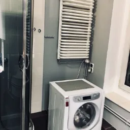 Wykończona łazienka z kabiną prysznicową, grzejnikiem drabinkowym i pralką Hotpoint Ariston, szare ściany, ciemna podłoga.