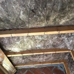 Ocieplenie poddasza wełną mineralną z widoczną konstrukcją drewnianą i siatką mocującą.