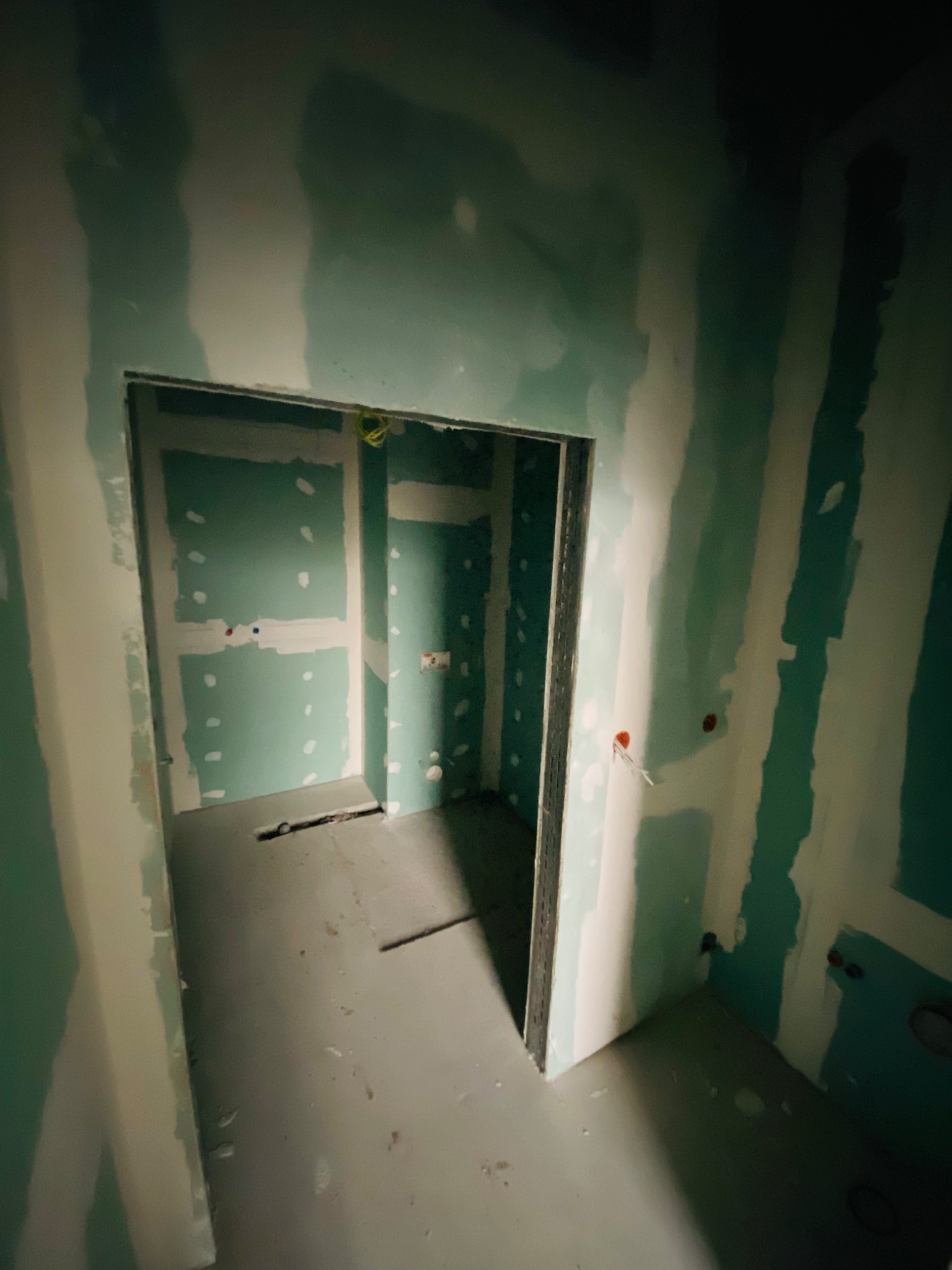 Wnętrze w trakcie prac wykończeniowych: ściany obłożone płytami gipsowo-kartonowymi, widoczne łączenia płyt wymagające dalszej obróbki, surowa posadzka, instalacja elektryczna częściowo wyprowadzona.