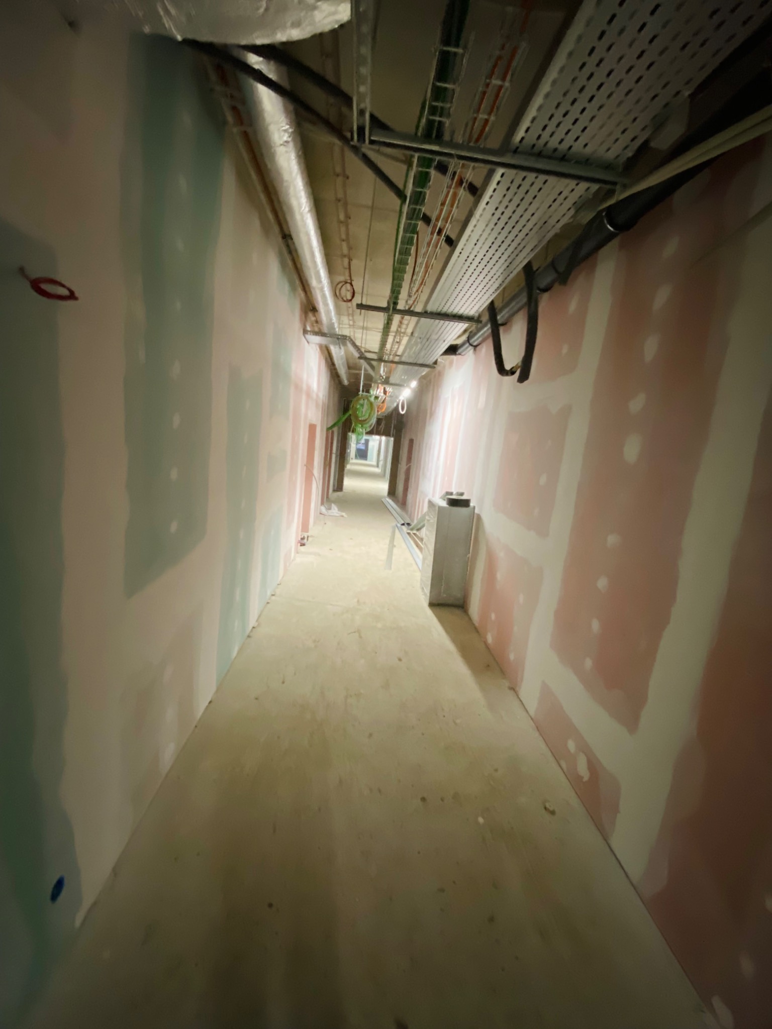 Długi korytarz w trakcie prac wykończeniowych: widoczne ściany pokryte płytami gipsowo-kartonowymi, wstępnie wyszpachlowane łączenia, instalacje pod sufitem.