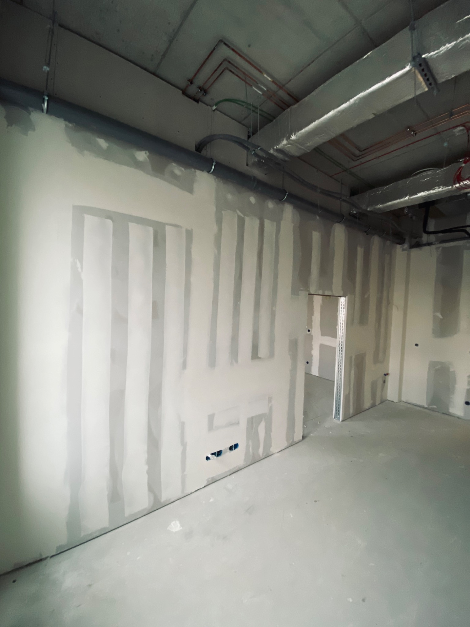 Wnętrze w trakcie prac wykończeniowych: ściany z płyt karton-gips, częściowo pokryte szpachlą, widoczne profile wzmacniające przy otworze drzwiowym, instalacje pod sufitem.