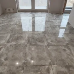 Ułożona, błyszcząca, szara podłoga z płytek w pokoju z oknami balkonowymi, widoczne odbicia światła na powierzchni płytek.