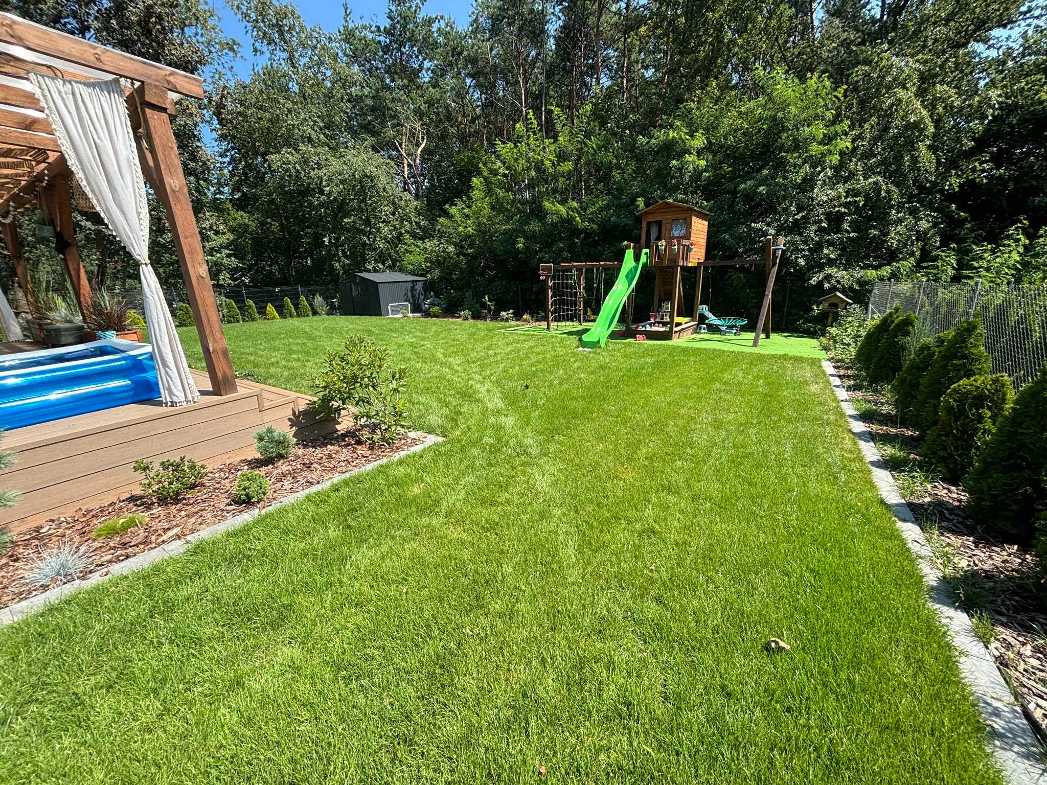 Zadbany trawnik z placem zabaw w ogrodzie. Widoczny basen, domek na drzewie ze zjeżdżalnią i huśtawkami. Trawa równo skoszona, obrzeża wykończone kostką. W tle drzewa.