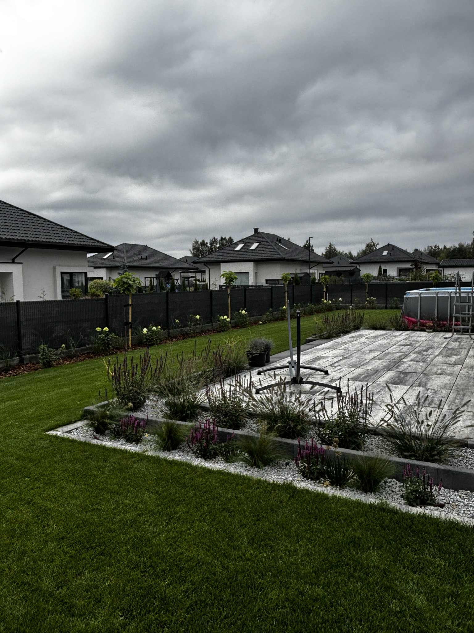 Nowoczesny ogród z równo przystrzyżonym trawnikiem, rabatami z ozdobnymi roślinami i kostką brukową w szarych tonacjach. W tle domy jednorodzinne i pochmurne niebo.
