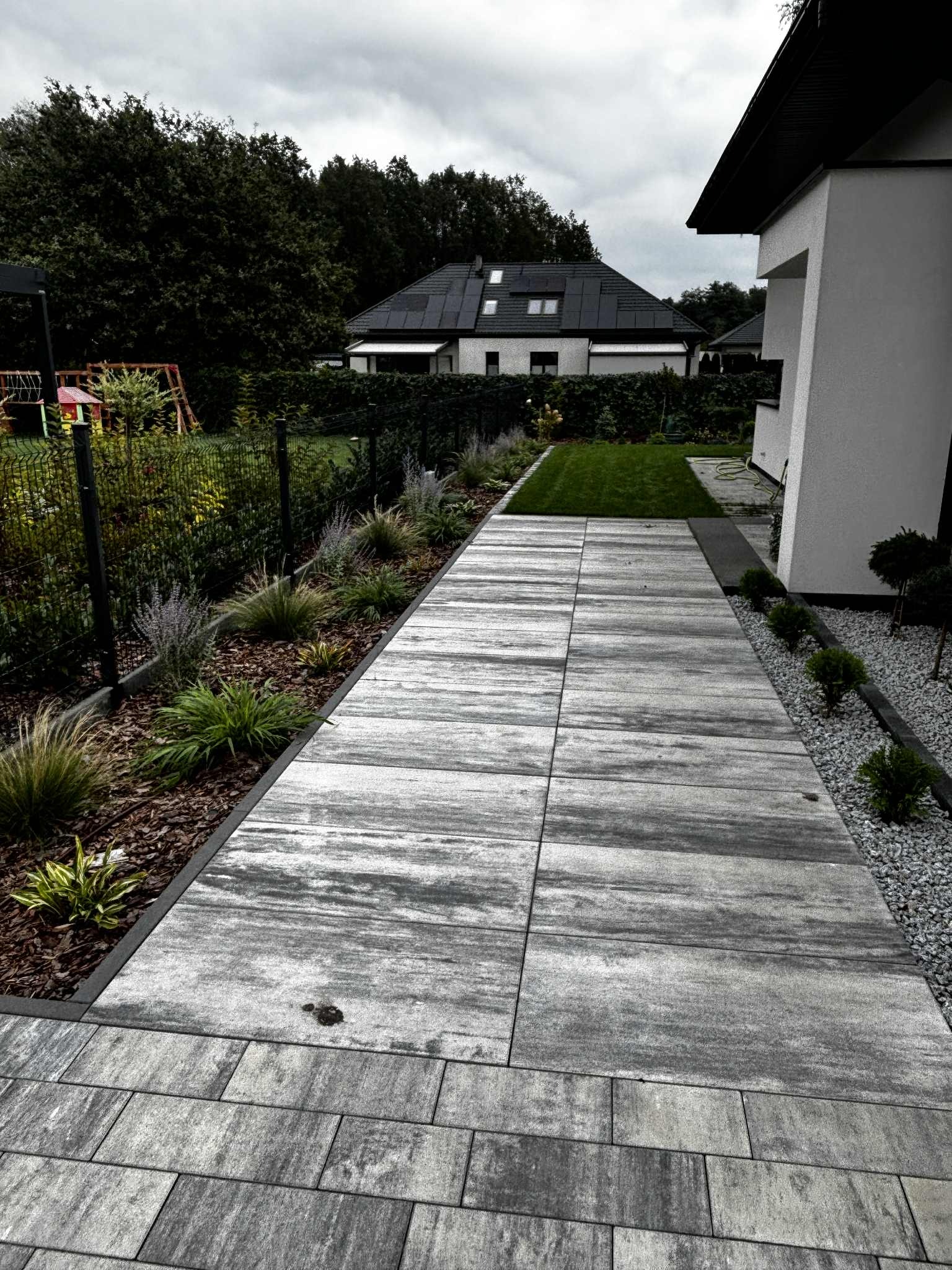 Ułożona szara kostka brukowa tworzy ścieżkę w ogrodzie z rabatami. Trawa z rolki, krzewy ozdobne i jasny żwir. W tle dom z panelami słonecznymi i plac zabaw.
