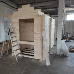 Wnętrze hali produkcyjnej w Pacółtowie z częściowo zmontowaną konstrukcją drewnianą, przypominającą saunę lub domek letniskowy, widoczna drabina i narzędzia.