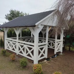 Biała, drewniana konstrukcja z czarnym dachem w ogrodzie, otoczona zielenią, z widocznymi elementami dekoracyjnymi i architektonicznymi.