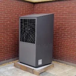 Szara pompa ciepła na drewnianej podstawie, umieszczona w rogu budynku z ceglaną ścianą, widok z przodu.