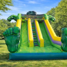 Nadmuchani - Kolorowa, dmuchana zjeżdżalnia w kształcie Hulka na trawie, z zielonymi rękami i stopami. Idealna atrakcja na imprezy plenerowe dla dzieci.