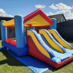 Kolorowy dmuchany zamek ze zjeżdżalniami na trawniku, widok z boku, błękitne niebo z chmurami w tle.