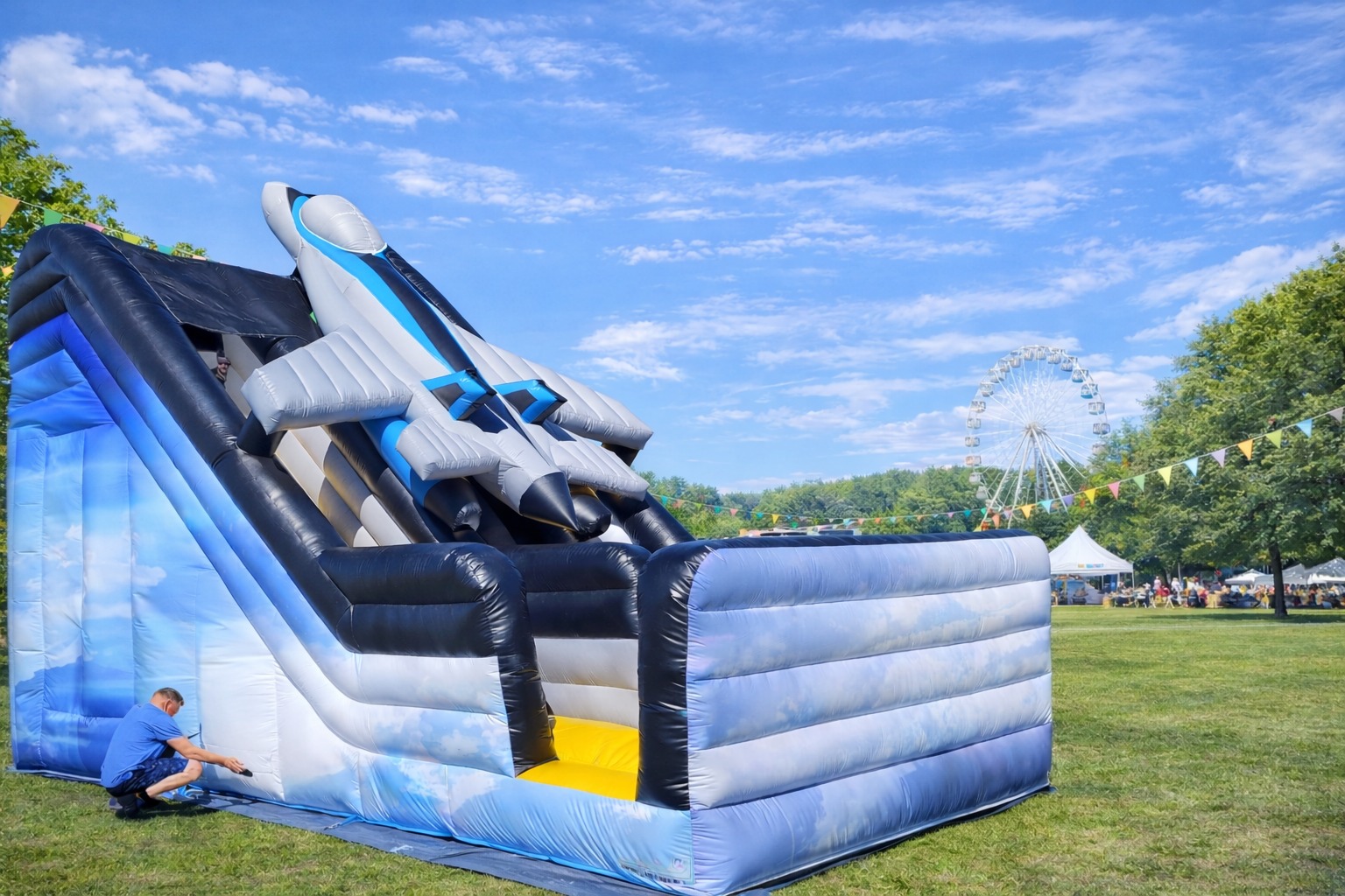 Rozłożona, duża, dmuchana zjeżdżalnia w kształcie samolotu na trawie, z mężczyzną przy podstawie i diabelskim młynem w tle, w słoneczny dzień.