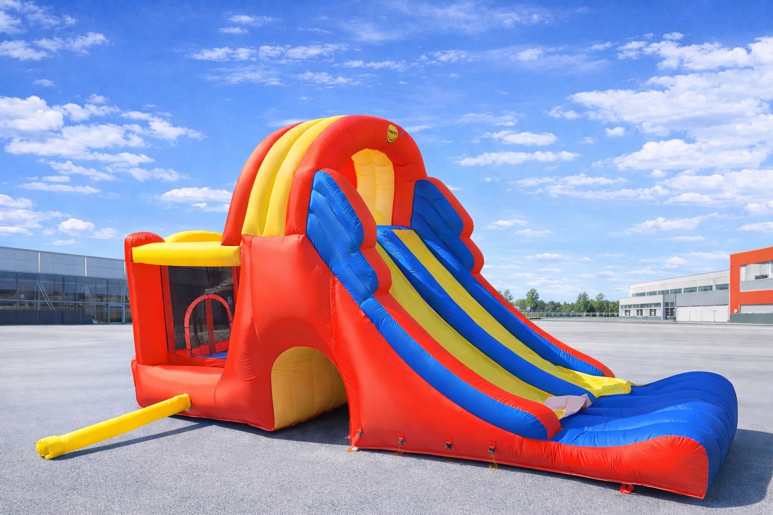 Kolorowa, dmuchana zjeżdżalnia z tunelem i basenem na szarym placu, pod błękitnym niebem. Widoczny budynek w tle. Zjeżdżalnia w odcieniach czerwieni, żółci i niebieskiego.