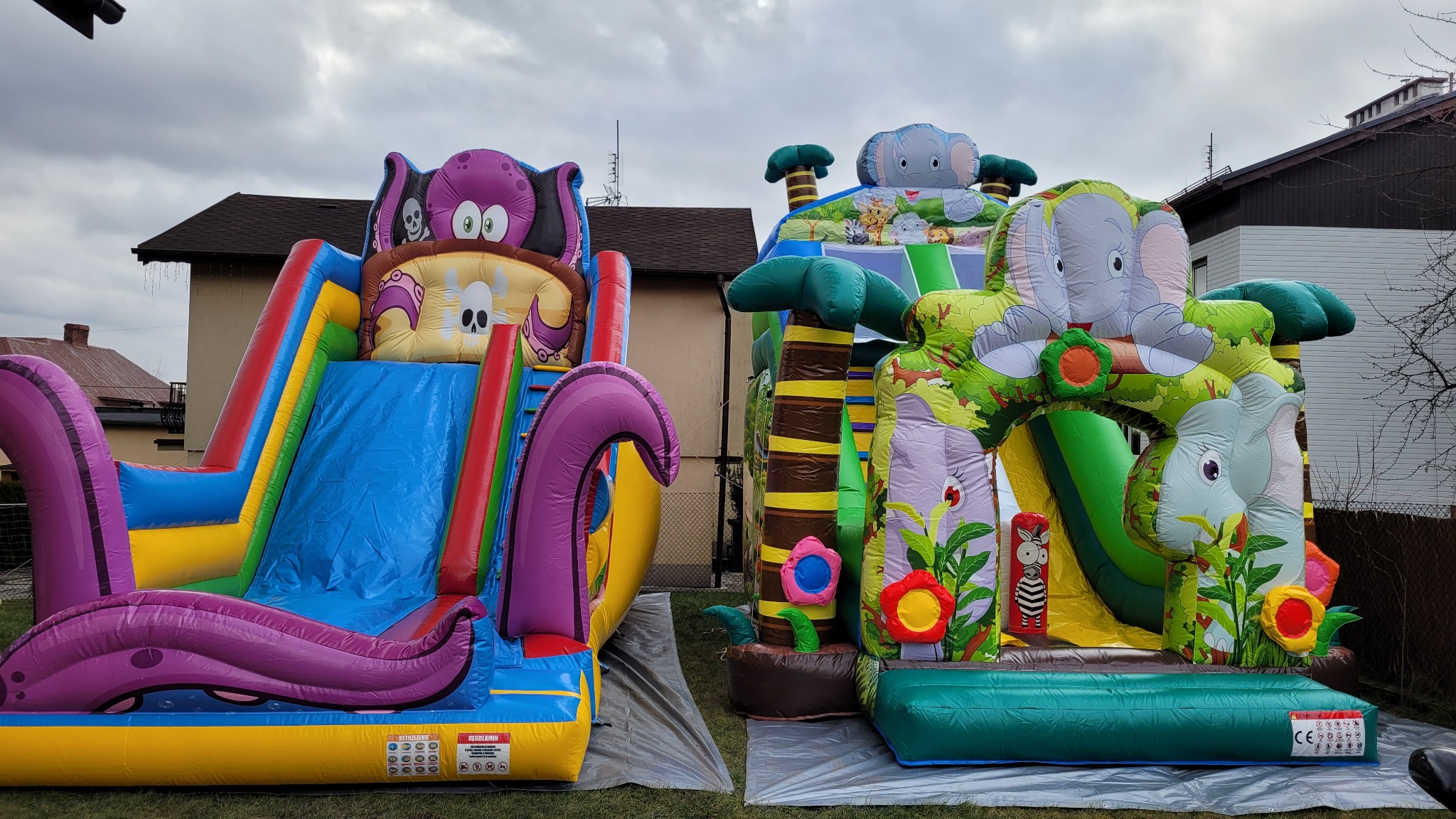 Dwa kolorowe dmuchańce: jeden z motywem ośmiornicy i czaszki, drugi z motywem słonia i dżungli, ustawione na trawie przed domem.