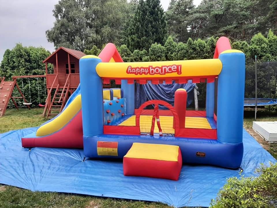 Kolorowy, dmuchany zamek ze zjeżdżalnią na niebieskiej plandece, w tle drewniany domek dla dzieci z huśtawkami.