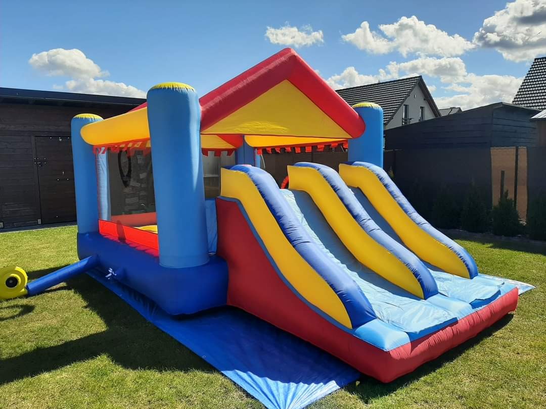 Kolorowy dmuchany zamek ze zjeżdżalniami na trawniku, widok z boku, błękitne niebo z chmurami w tle.