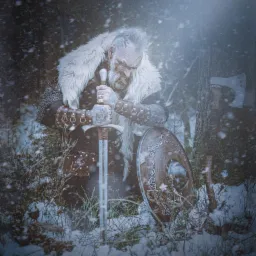 Mężczyzna w kostiumie wikinga klęczy w śniegu, trzymając miecz, obok tarczy i topora.