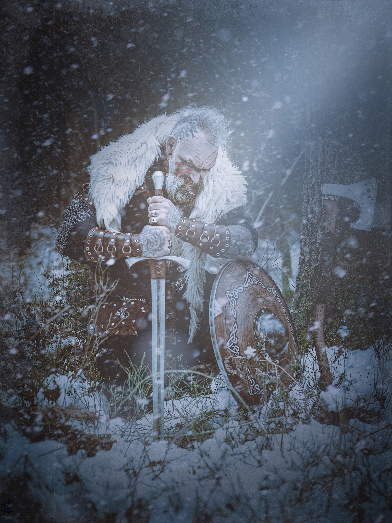 Mężczyzna w kostiumie wikinga klęczy w śniegu, trzymając miecz, obok tarczy i topora.