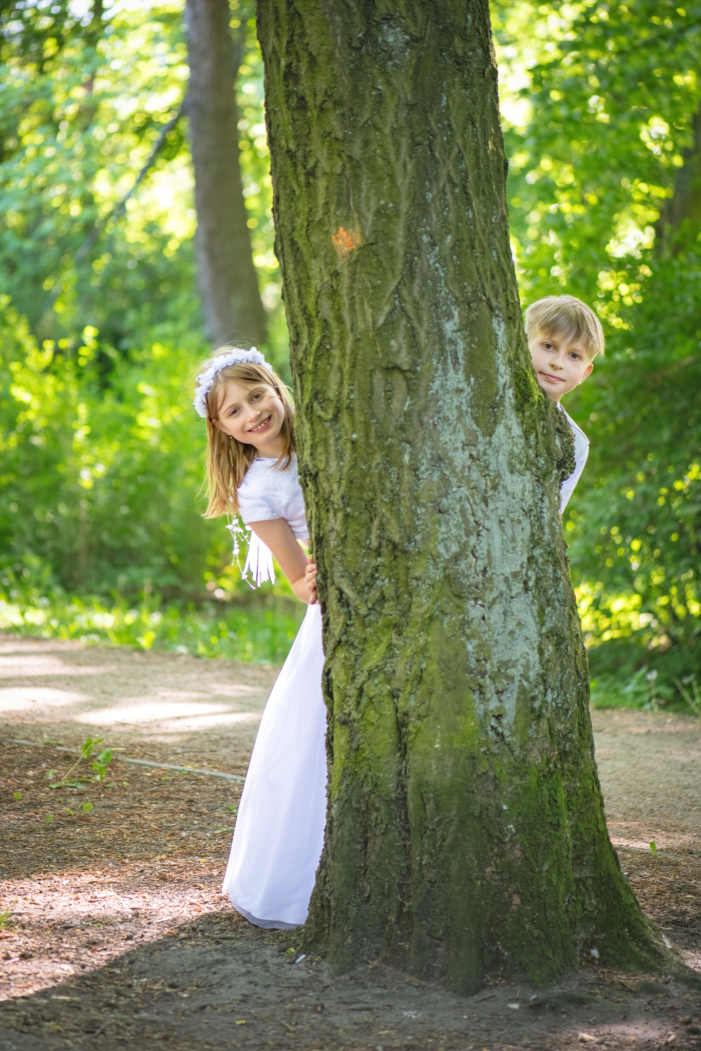 Dziewczynka w białej sukience komunijnej i chłopiec w białej koszuli chowają się za grubym, omszałym drzewem w lesie, patrząc w obiektyw.
