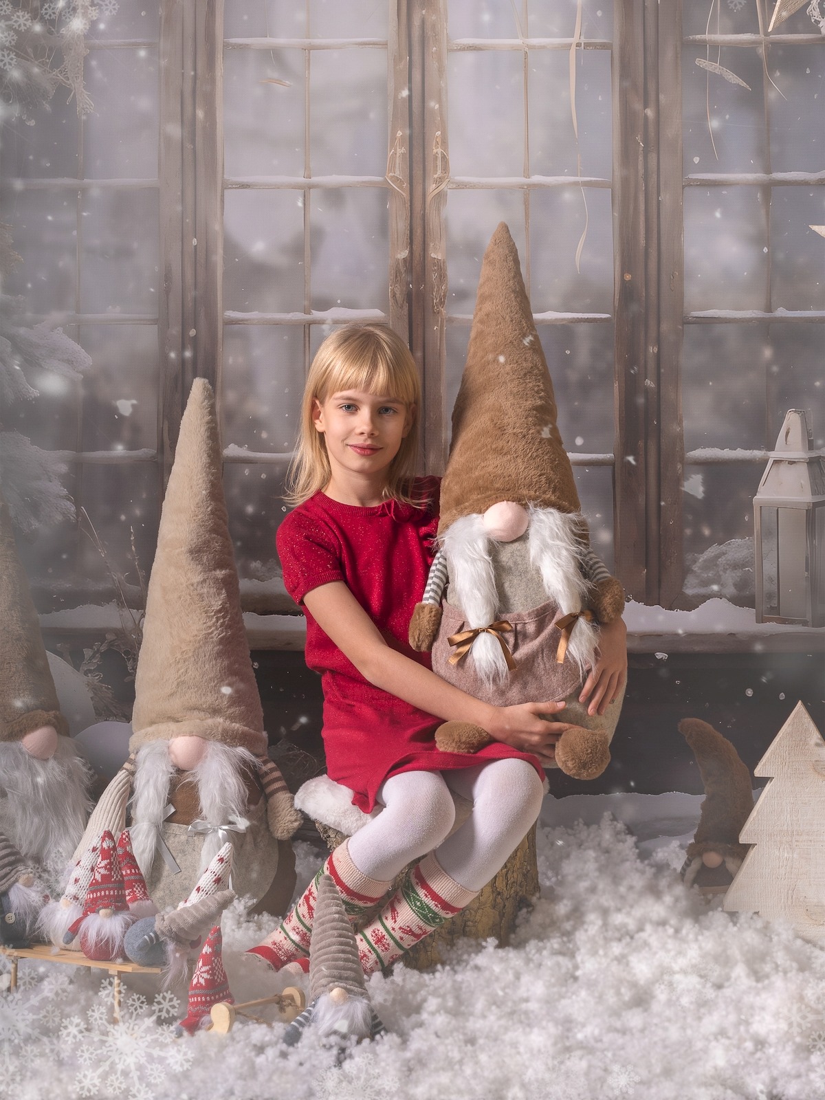 Dziewczynka w czerwonej sukience siedzi w świątecznej scenerii, trzymając dużego, pluszowego gnoma na tle okna i dekoracji zimowych.