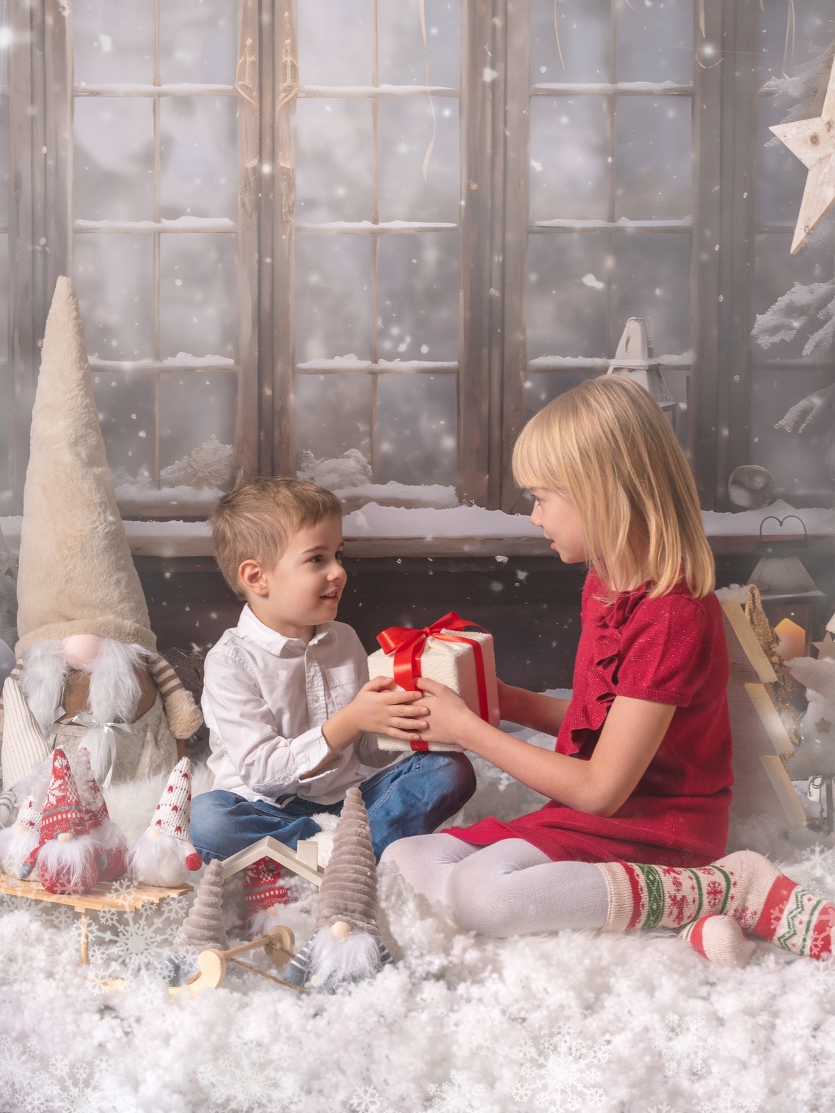 Świąteczna sesja: Dziewczynka w czerwonej sukience wręcza prezent chłopcu na tle okna i dekoracji bożonarodzeniowych, w otoczeniu pluszowych skrzatów i sztucznego śniegu.