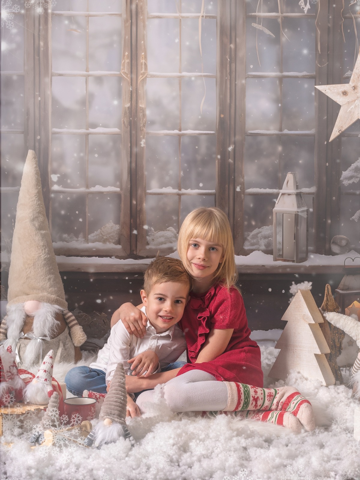 Uśmiechnięte rodzeństwo, brat i siostra, w świątecznej scenerii z dekoracjami bożonarodzeniowymi i sztucznym śniegiem, pozują na tle okna. Dziewczynka w czerwonej sukience.