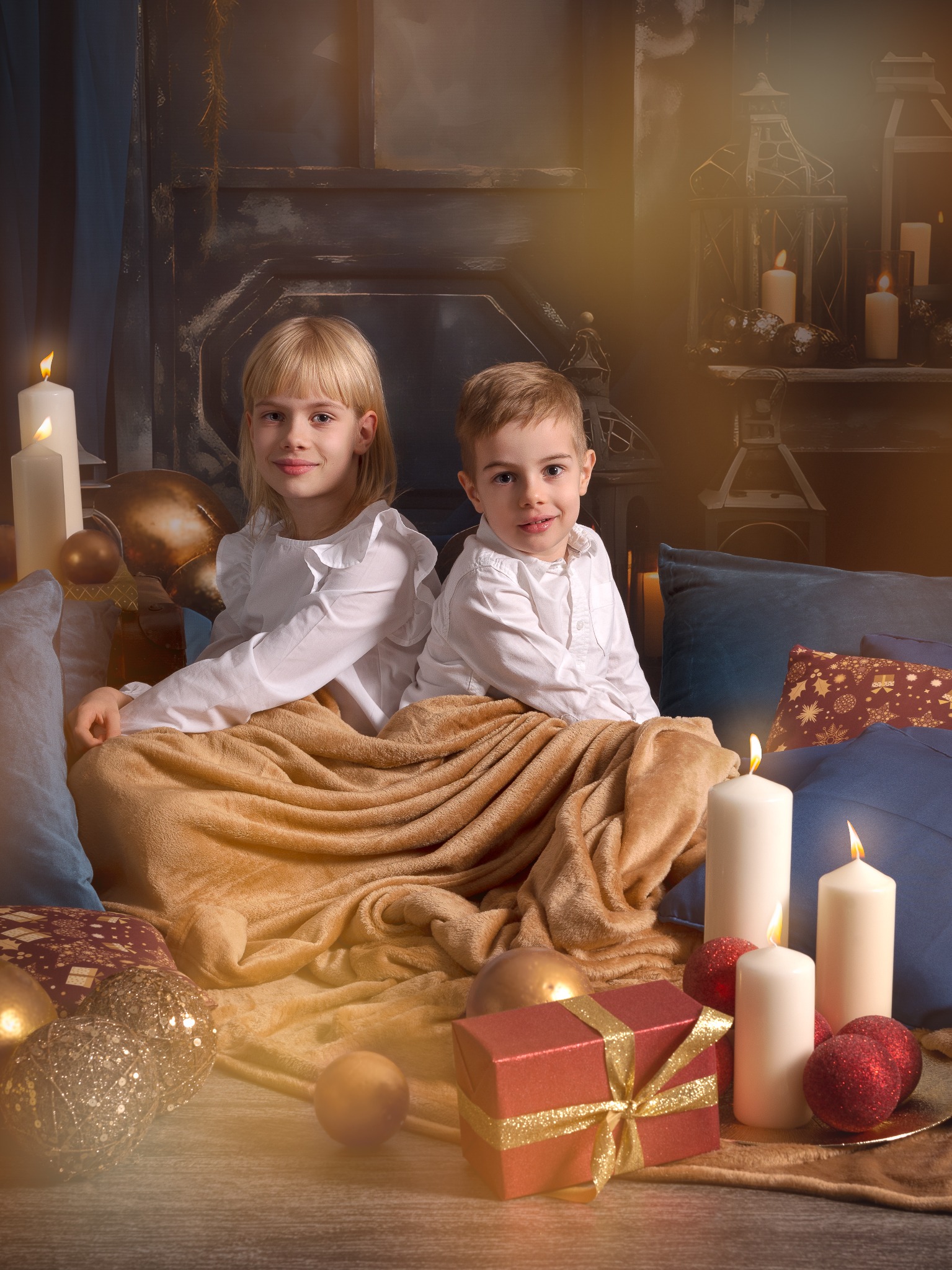 Uśmiechnięte rodzeństwo, blond chłopiec i dziewczynka, w białych koszulach, otuleni kocem na tle świątecznej scenografii z prezentami i świecami, w ciepłym oświetleniu.