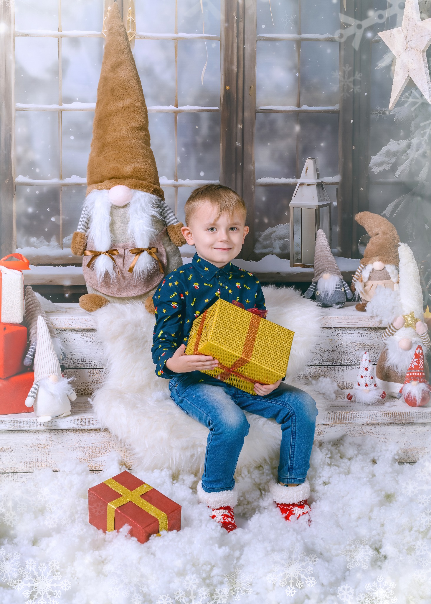 Chłopiec w świątecznej scenerii siedzi na futrze z prezentem, otoczony skrzatami i dekoracjami. W tle okno z zimowym krajobrazem. Sesja świąteczna Boże Narodzenie.