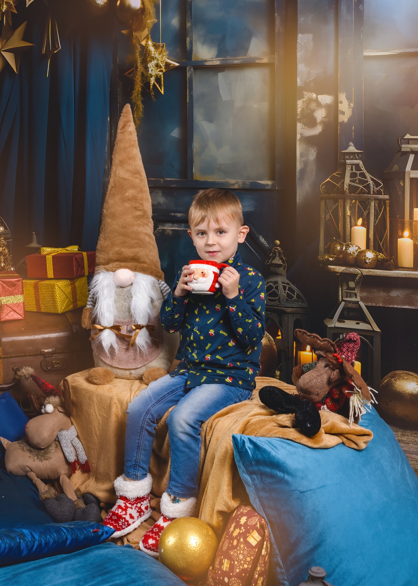 Chłopiec z blond czupryną pije z kubka w świątecznej scenerii z pluszowym skrzatem, reniferem i prezentami w tle, tworząc ciepłą, rodzinną atmosferę.