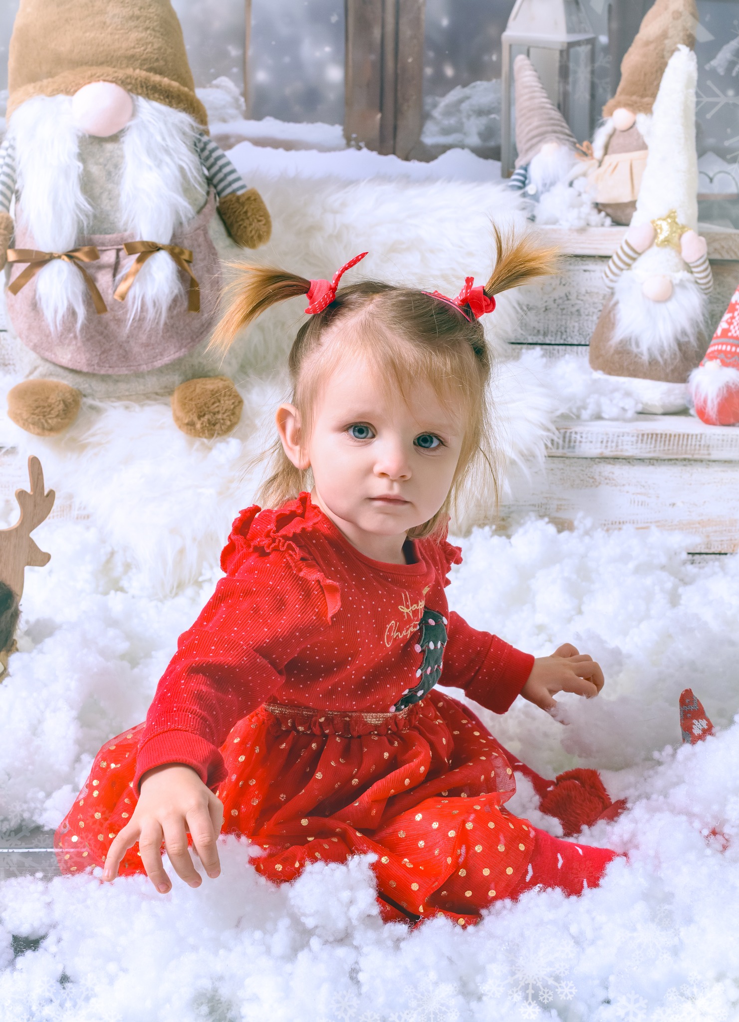Uśmiechnięta dziewczynka w czerwonej sukience siedzi na sztucznym śniegu, otoczona świątecznymi dekoracjami i gnomami. Scena z sesji bożonarodzeniowej.