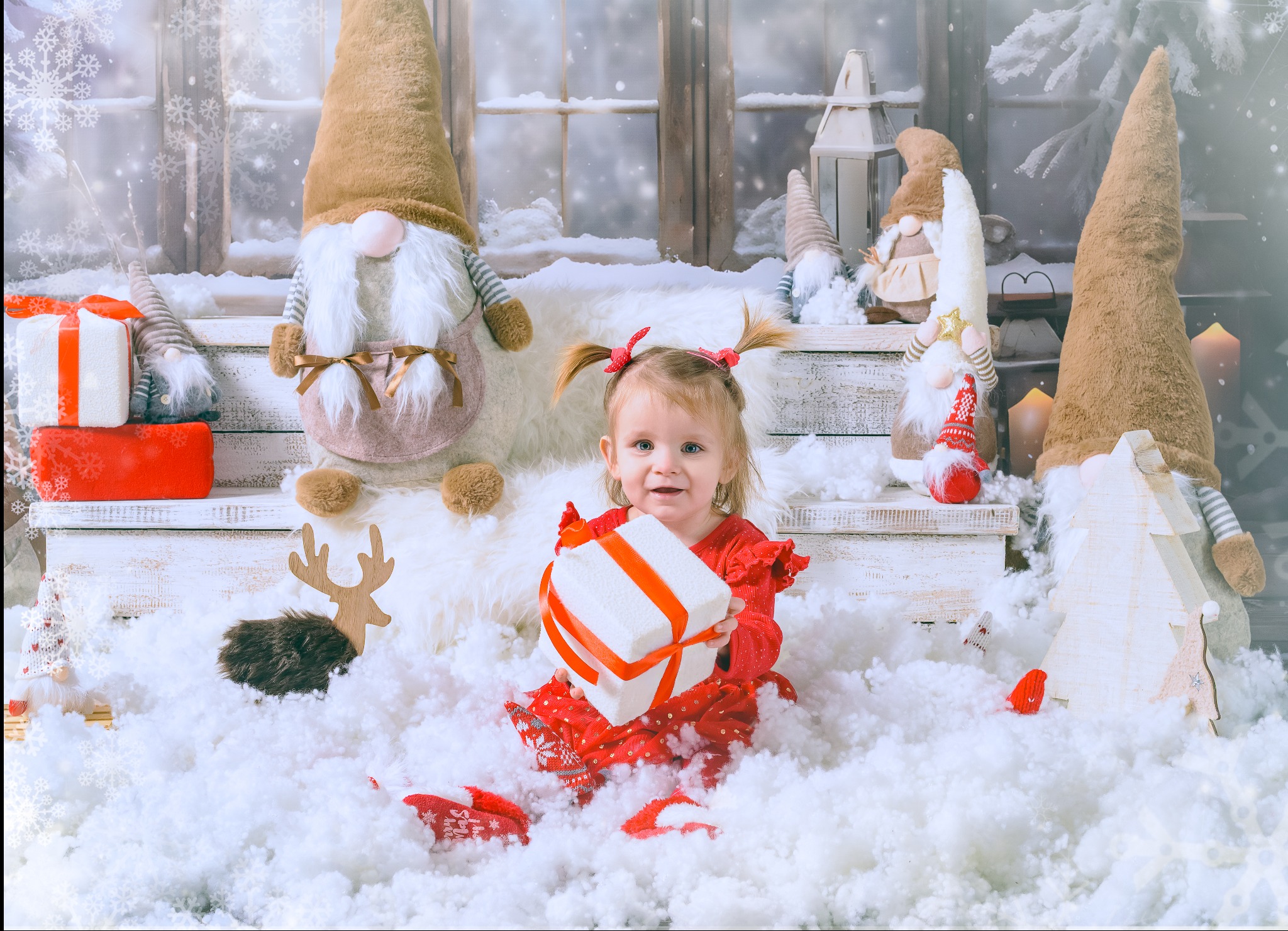 Uśmiechnięta dziewczynka w czerwonej sukience siedzi w sztucznym śniegu, trzymając prezent na tle świątecznej scenografii z gnomami i oknem.