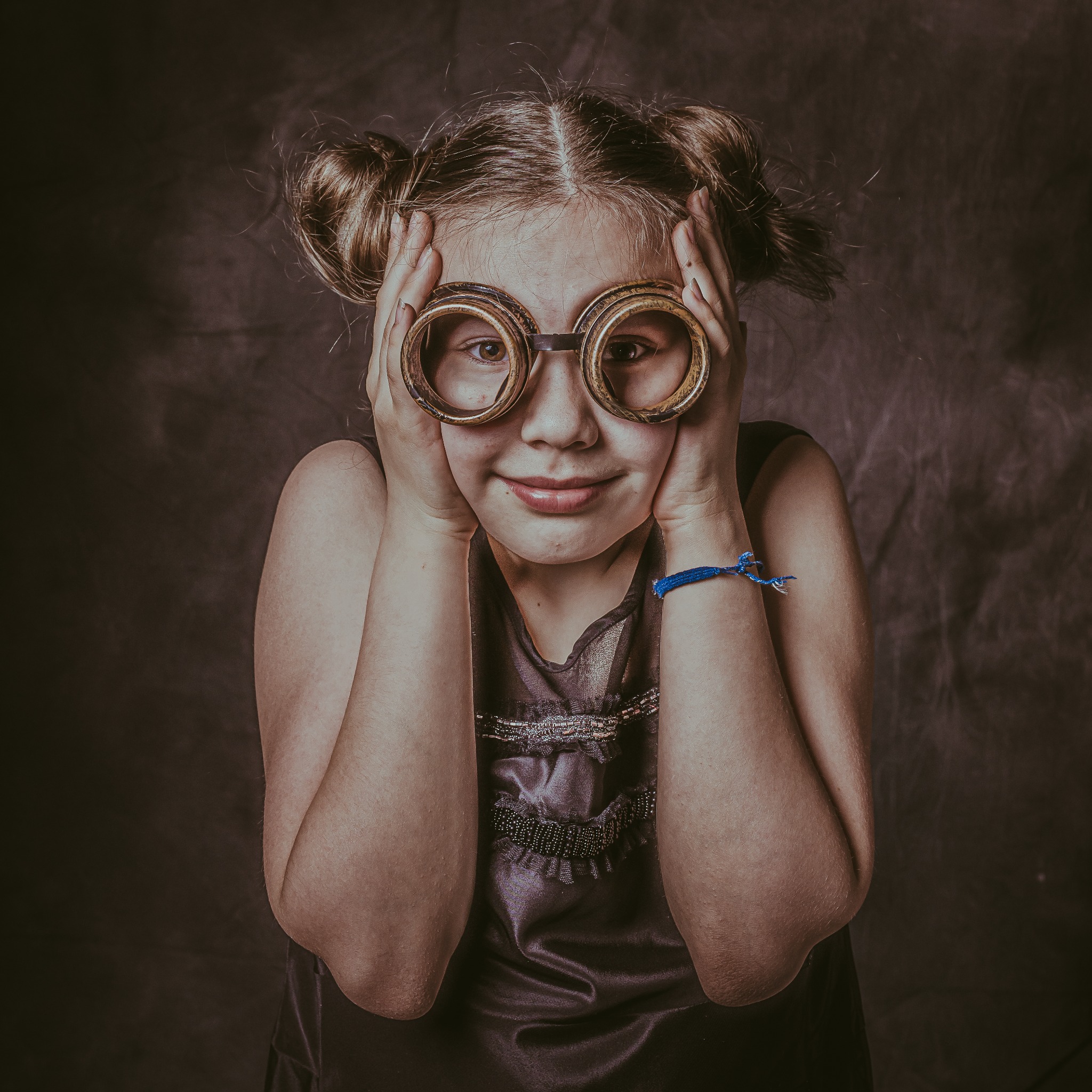 Portret uśmiechniętej dziewczynki w dużych, okrągłych okularach w stylu retro, z włosami upiętymi w dwa koczki i niebieską bransoletką na nadgarstku, na ciemnym, rozmytym tle.