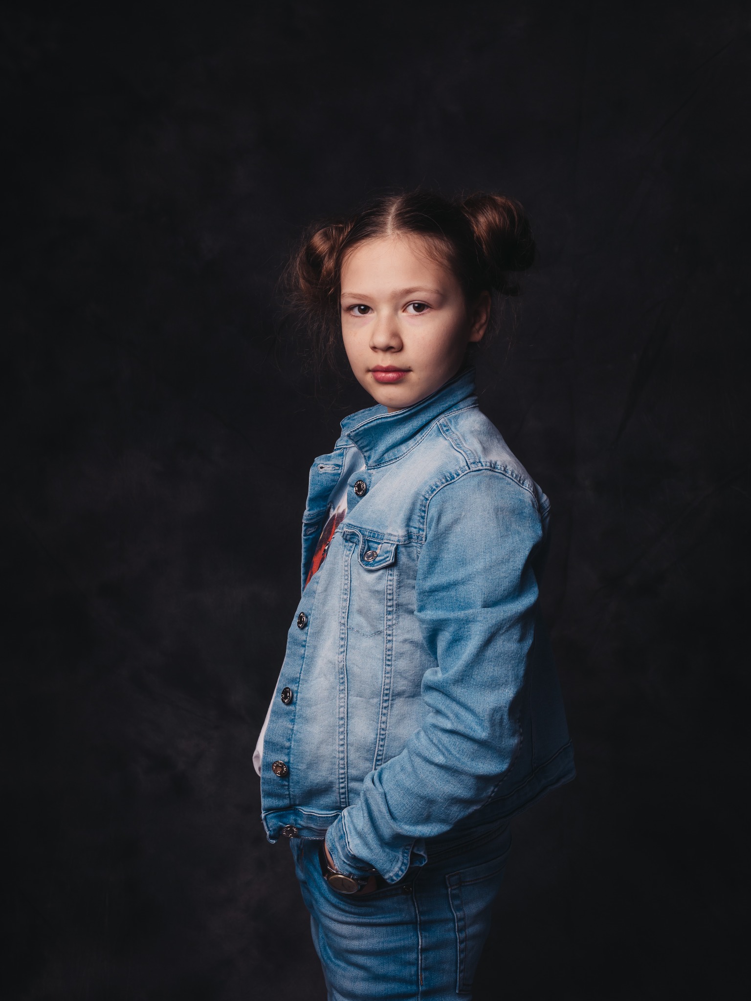 Portret dziewczynki w dżinsowej kurtce, z włosami upiętymi w dwa koczki, pozującej na ciemnoszarym tle. Dziewczynka ma rękę w kieszeni i patrzy wprost w obiektyw.