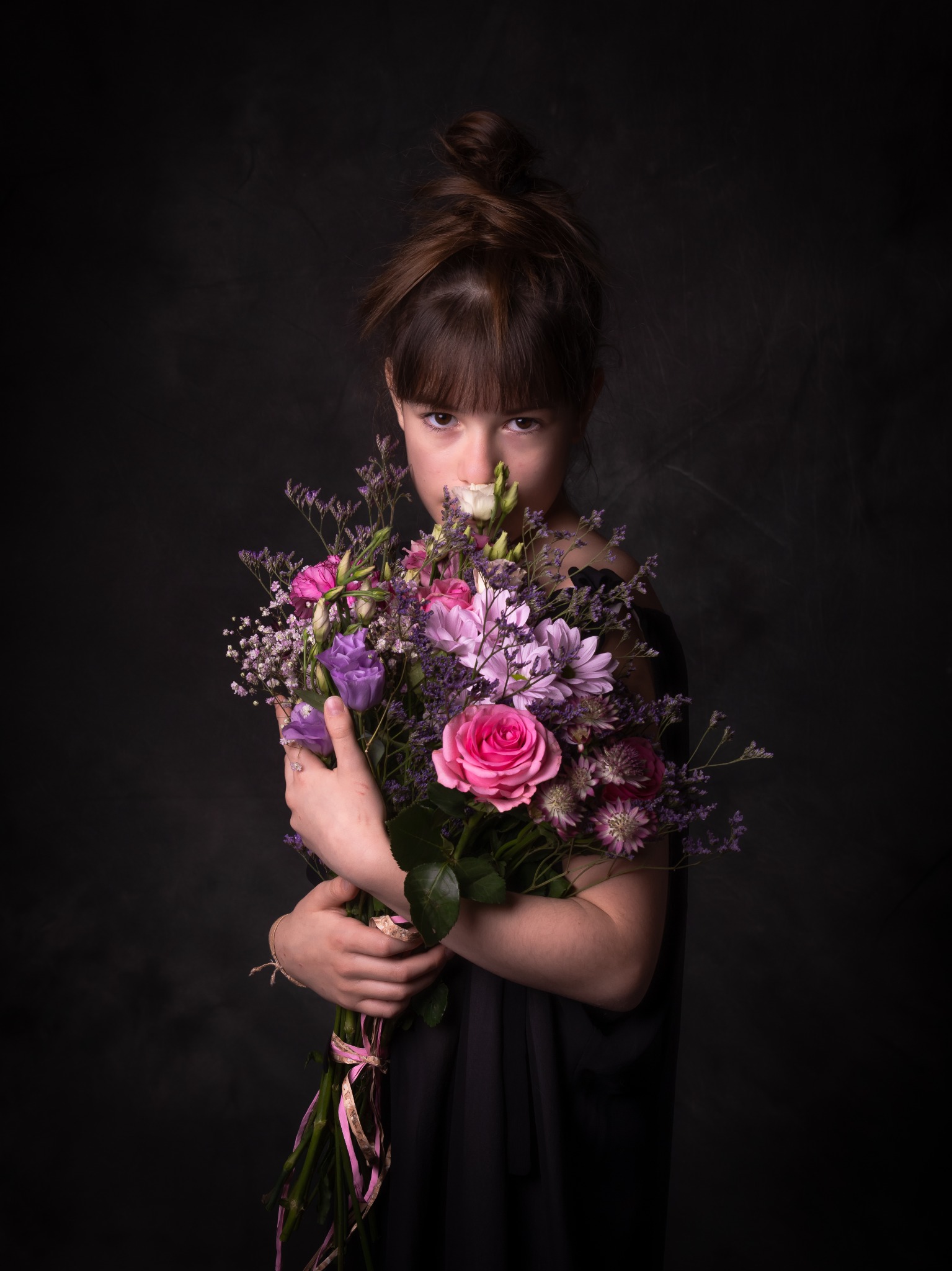 Dziewczynka z brązowymi włosami upiętymi w kok, trzymająca duży bukiet różowych, fioletowych i białych kwiatów na ciemnym, rozmytym tle.