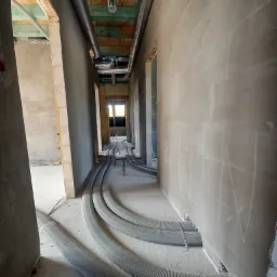 Wnętrze budynku w trakcie montażu instalacji wentylacyjnej: widoczne szare, karbowane rury wentylacyjne prowadzone po podłodze w surowym, otynkowanym korytarzu. Na ścianach widoczne pomocnicze...