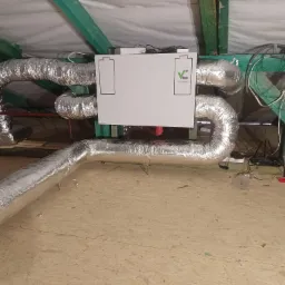 Szare, elastyczne przewody wentylacyjne podłączone do centrali wentylacyjnej z logo Vent Clear, zamontowane na poddaszu z drewnianą konstrukcją nośną.