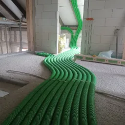 Wiązka zielonych, karbowanych rur wentylacyjnych ułożona na szorstkiej, betonowej posadzce w surowym wnętrzu budynku w trakcie budowy. Rury prowadzą przez otwór w ścianie z betonu komórkowego.