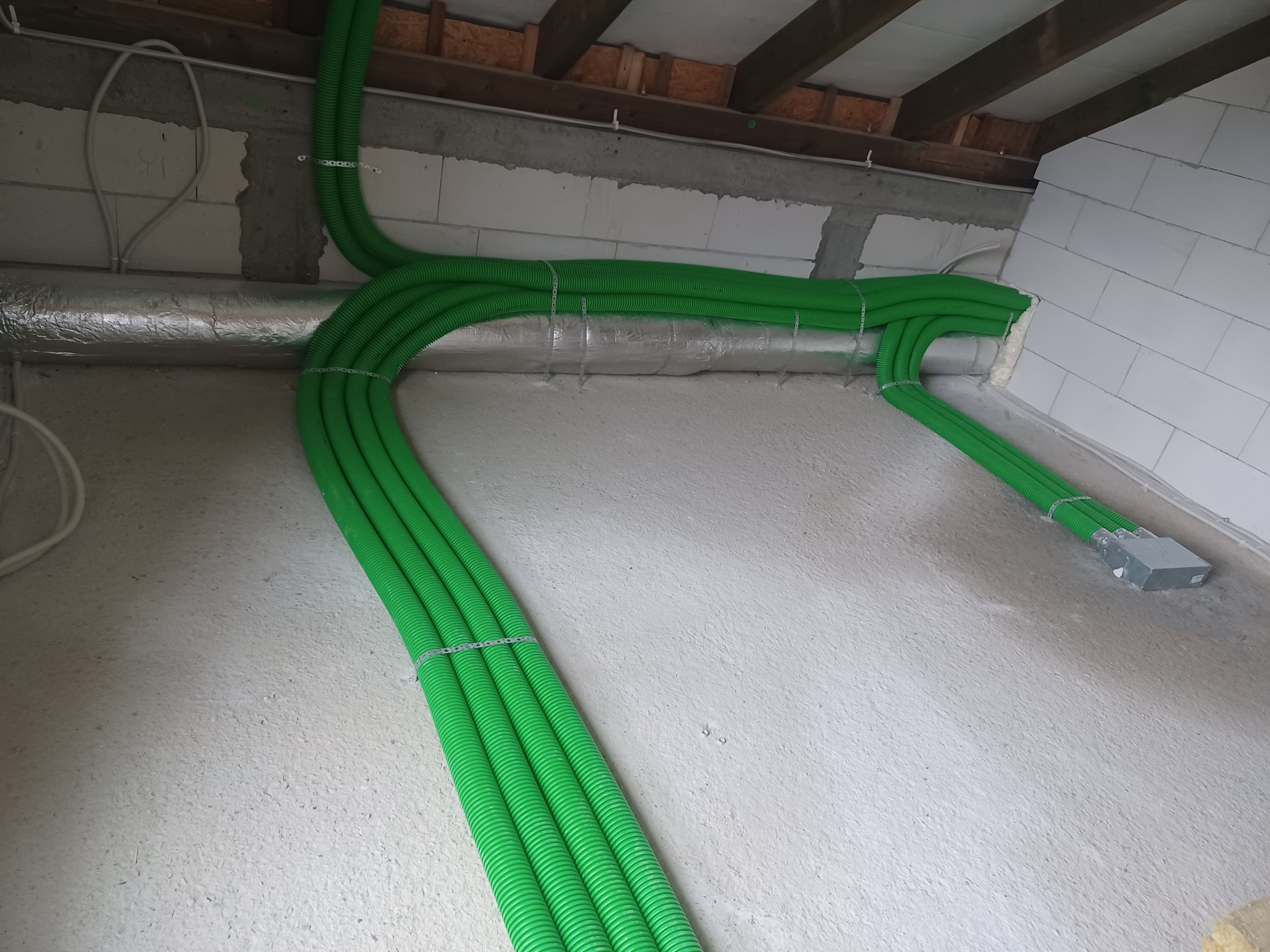 Zielone, karbowane rury wentylacyjne prowadzone pod sufitem w surowym wnętrzu, spięte metalowymi obejmami, z widoczną izolacją termiczną na rurze i betonową ścianą w tle.