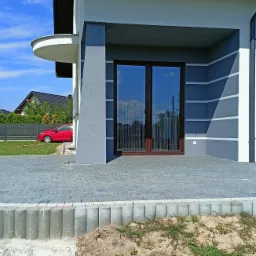 Szare ocieplenie domu z dekoracyjnymi białymi pasami, widoczne dwuskrzydłowe drzwi balkonowe z brązowymi ramami i szara kostka brukowa na tarasie.