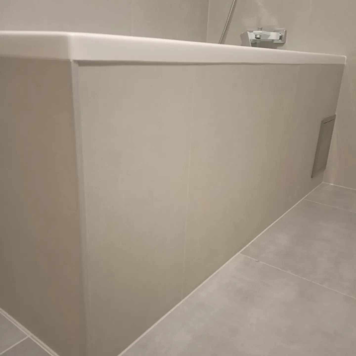 Obudowana wanna z szarymi płytkami, widoczny fragment baterii i rewizja.