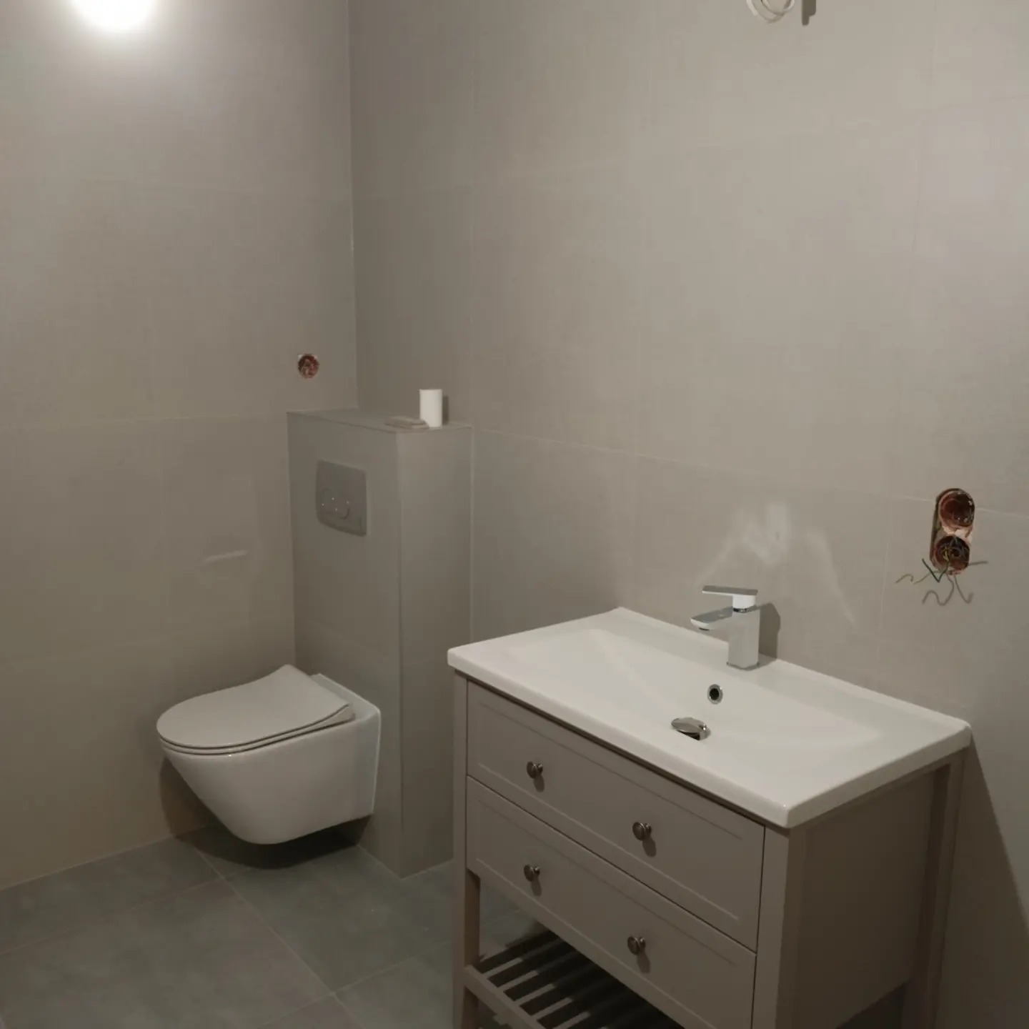 Szara łazienka w Bielsku-Białej z podwieszaną toaletą i szafką pod umywalkę z szufladami, widoczne niedokończone podłączenie elektryczne.