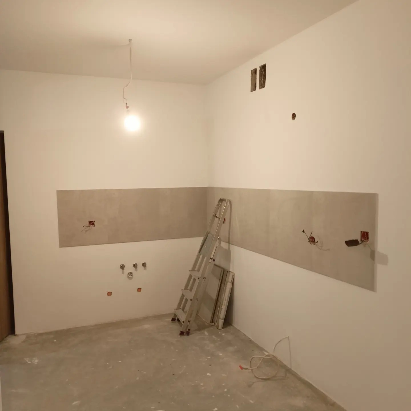 Wnętrze w trakcie prac wykończeniowych: częściowo położone płytki ścienne, widoczne instalacje elektryczne i hydrauliczne, aluminiowa drabina oparta o ścianę.