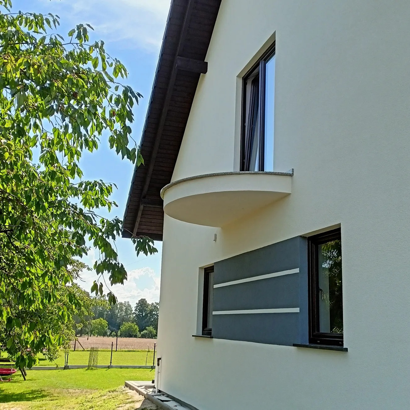 Elewacja domu jednorodzinnego z oknami, balkonem i szarym panelem dekoracyjnym, widok z boku na tle zieleni i błękitnego nieba.