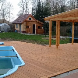 Nowoczesny taras z drewna z basenem i zadaszeniem, widok na domek letniskowy i trampolinę w ogrodzie.