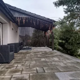 Zadaszony taras wyłożony szarą kostką brukową, z czarnymi meblami ogrodowymi i dekoracyjnym oświetleniem w postaci girlandy żarówek.