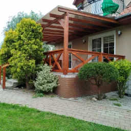 Drewniana pergola z ażurową balustradą, przylegająca do domu z balkonem. Widoczne rośliny ozdobne, ścieżka z kostki i zbiornik na deszczówkę.