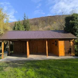 Drewniana konstrukcja z brązowym dachem na betonowej kostce, otoczona zielenią i drzewami, w tle wzgórze pod błękitnym niebem. Widoczna część zadaszona i zamknięta z drzwiami.