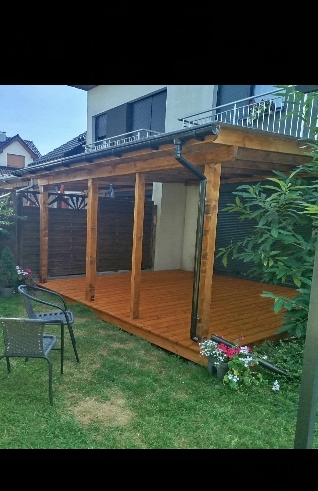 Zadaszona konstrukcja z drewnianych belek i desek, tworząca otwartą przestrzeń na zewnątrz domu, z widocznym fragmentem brązowego ogrodzenia w tle i dwoma krzesłami ogrodowymi na trawniku.