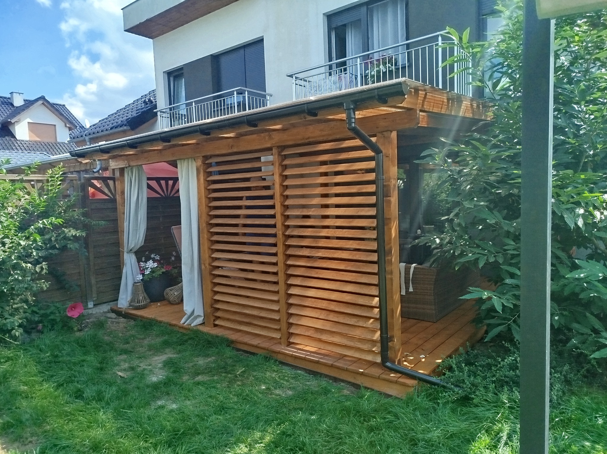 Drewniana konstrukcja z żaluzjowymi ścianami i zasłonami, przylegająca do domu z balkonem, na trawniku z kwiatem hibiskusa.